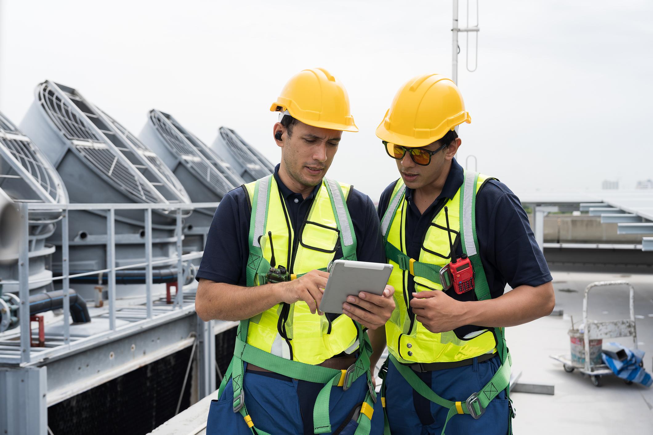 Two maintenance technicians in hi-vis vests and hard hats reviewing asset condition data on a tablet beside rooftop cooling plant